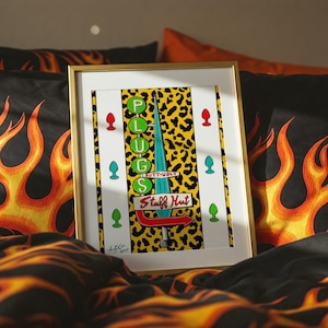 May include: A framed artwork with a gold-colored frame. The artwork features the word "PLUGS" vertically, a sign that says "BUTT-UGLY Stuff Hut", and several colorful plug illustrations. The background has a yellow leopard print.