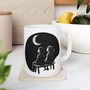 May include: White ceramic mug featuring a black and white illustration of two figures seated on a bench, gazing at a crescent moon and stars in a night sky. The mug has a curved handle.