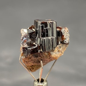 Op de afbeelding: Een donker, glanzend zwart mineraal specimen met een kristallijne structuur. Het mineraal is gemonteerd op een metalen standaard, die de geometrische vorm en natuurlijke schoonheid laat zien. Het specimen heeft een ruwe, bruine matrix.