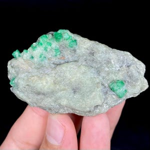 May include: A rough, irregularly shaped rock with several vibrant green emerald crystals embedded within. The stone has a light gray and white base color, with a few small green crystals scattered across its surface. The background is black.
