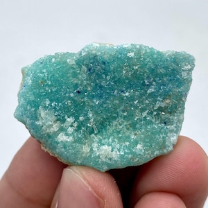 Puede incluir: Primer plano de un espécimen mineral en bruto. La piedra es de un color turquesa vibrante con parches de azul más oscuro y pequeñas formaciones de cristales blancos. El mineral tiene una superficie texturizada e irregular.
