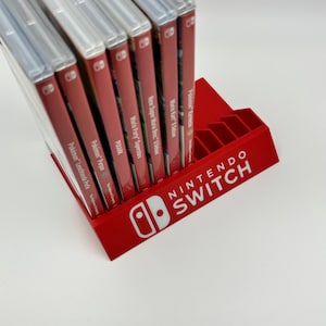 Könnte beinhalten: Ein roter Nintendo Switch-Spielkassettenhalter mit dem Nintendo Switch-Logo in Weiß. Der Halter ist mit mehreren Spielhüllen gefüllt, darunter "Pokémon Leuchtende Perle", "Pokémon Purpur", "Mario Party Superstars" und "Mario Kart 8 Deluxe".