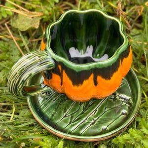 Könnte beinhalten: Ein Keramik-Tassen- und Untertassen-Set mit Kürbis-Design. Die Tasse ist orange und grün mit einem schwarzen Streifen, und die Untertasse ist grün mit einem Blättermuster. Der Griff der Tasse ist grün gestreift.