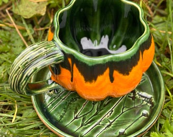 May include: A ceramic cup and saucer set with a pumpkin design. The cup is orange and green with a black stripe, and the saucer is green with a leaf pattern. The handle of the cup is striped green.