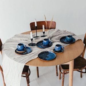 May include: A round wooden dining table set with a light beige linen tablecloth, featuring blue ceramic plates, cups, and saucers. Black candle holders with white candles and wooden chairs complete the dining set.