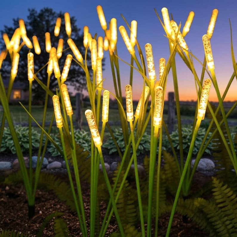 Pu&ograve; includere: Una collezione di luci da giardino per esterni a forma di alti steli d'erba con punte cilindriche testurizzate e luminose. Ogni stelo presenta un sottile gambo verde che sostiene un elemento illuminante caldo che emette un bagliore dorato.