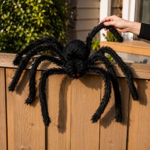 May include: A large, fuzzy black spider decoration featuring eight long, posable legs covered in synthetic fur. The spider has a round, textured body and two small, glowing red eyes positioned at the front, designed for Halloween display.