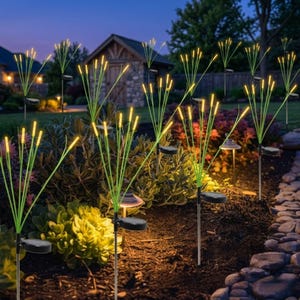 Pu&ograve; includere: Una serie di luci solari da giardino con sottili steli verdi sormontati da lampadine gialle luminose. Ogni luce include un piccolo pannello solare circolare alla base, disposte in un'aiuola con terreno scuro e varie piante verdi.