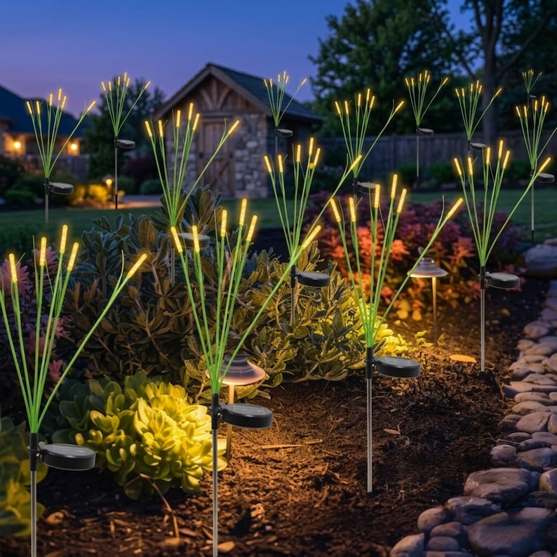 Pu&ograve; includere: Una serie di luci solari da giardino con sottili steli verdi sormontati da lampadine gialle luminose. Ogni luce include un piccolo pannello solare circolare alla base, disposte in un'aiuola con terreno scuro e varie piante verdi.
