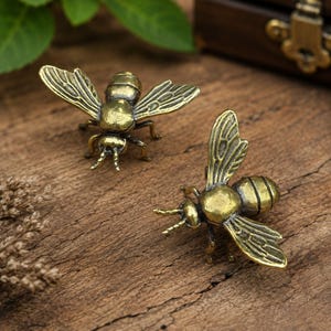 May include: Two small, three-dimensional bee figurines crafted from a metallic material with an antique brass finish. Each bee features detailed wings with etched vein patterns, a segmented body, and small antennae, capturing a realistic insect form.