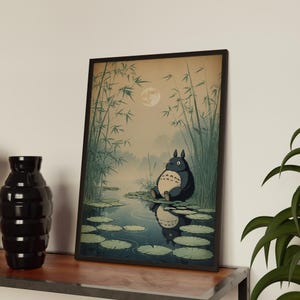 May include: Framed art print depicting a tranquil scene with a large, dark blue creature, similar to Totoro, sitting by a pond with lily pads. The artwork includes bamboo, a full moon, and a reflection in the water.