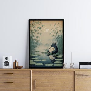 May include: Framed art print depicting a tranquil scene with a large, dark grey creature, similar to Totoro, seated on lily pads in a pond. The background features bamboo, a full moon, and a misty atmosphere, creating a peaceful, nature-themed artwork.
