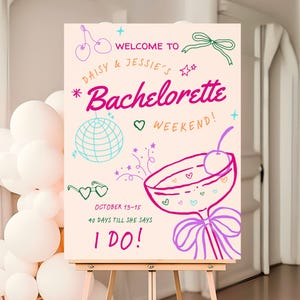 Könnte beinhalten: Ein hellrosa Schild auf einer hölzernen Staffelei mit den Worten "Welcome to Daisy & Jessie's Bachelorette Weekend!" in einer skurrilen Schriftart. Das Schild enthält Illustrationen von Kirschen, einer Discokugel und einem Cocktail.