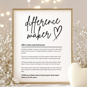 May include: A printable wall art print with a black and white design. The print features the word "difference maker" with a heart symbol and a definition of the term. The print is framed in a gold frame and is sitting on a white surface.