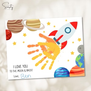 May include: A white card with a rocket ship, planets, and stars. The card features a handprint in orange and yellow, with the text "I LOVE YOU TO THE MOON & BACK!" and a handwritten signature.