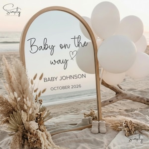 Op de afbeelding: Een houten spiegel met de tekst "Baby on the way" en "Baby Johnson October 2026". De spiegel is omgeven door witte ballonnen, droogbloemen en babyschoentjes, op een strand.