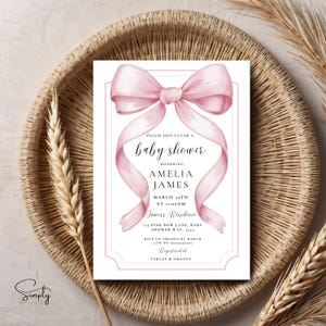 May include: Pink baby shower invitation with a watercolor illustration of a pink bow. The invitation text reads "Please join us for a baby shower honoring Amelia James, March 20th at 11:00am, Jamess Residence, Pink Bow Lane, Baby Shower Way, 5555, RSVP to Amanda by March 14th on, Registered at Target & Amazon."