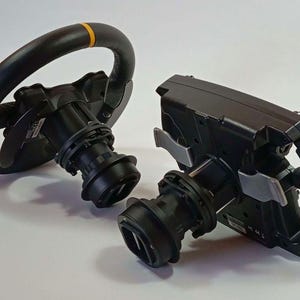 May include: Two black racing steering wheels for gaming simulation. One wheel is circular with a yellow top marker, while the other is a flat-bottomed rectangular style. Both feature central mounting hubs, paddle shifters, and textured grips.