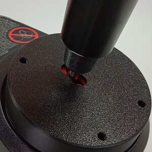 Könnte beinhalten: Ein schwarzer Joystick-Controller mit einer texturierten kreisförmigen Basis, vier Schraubenlöchern und einem zentralen roten Drehpunkt. Ein Warnetikett ist an der Seite sichtbar, das einen roten Kreis mit Schrägstrich über einem Handsymbol zeigt.