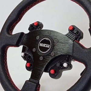 May include: A black racing steering wheel with red stitching on the grip. The center features a brushed metal plate with the Sparco logo, carbon fiber accents, and multiple red and black buttons mounted on the spokes for control inputs.