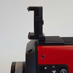Op de afbeelding: Een rood en zwart elektronisch apparaat met een verticale zwarte smartphonehouder aan de bovenkant. Het apparaat heeft een zwart bedieningspaneel met een wit pijlpictogram en een wit vierkant pictogram met de tekst STOP en een klein icoon.