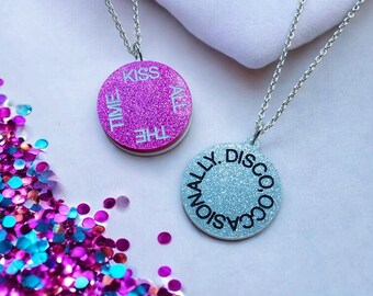 May include: Two circular pendant necklaces with silver chains. One is glittery pink with the words "Kiss All The Time". The other is glittery blue with the words "Occasionally Disco".