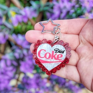 May include: A Diet Coke heart-shaped keychain with a red glitter border and a silver star-shaped clasp. The keychain features the Diet Coke logo in red and white.