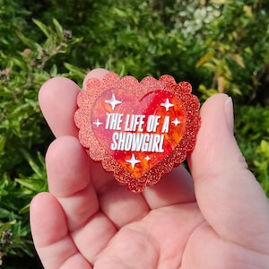 Pode incluir: Um broche em forma de coração com uma borda vermelha brilhante e o texto "THE LIFE OF A SHOWGIRL" em branco. O broche tem um design marmorizado vermelho e laranja no centro, com quatro estrelas brancas.