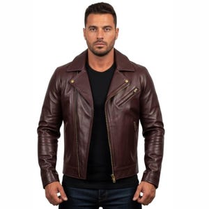 Puede incluir: Chaqueta de cuero burdeos con un estilo motero clásico. La chaqueta presenta un cuello con muescas, cierre de cremallera asimétrico y múltiples bolsillos con cremallera. El modelo lleva una camiseta negra y vaqueros azules.