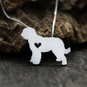 Peut inclure: Collier en argent avec une silhouette découpée d'un chien doodle avec un petit cœur au centre. Le collier est posé sur une surface en bois.