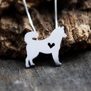 May include: A silver-colored pendant necklace featuring a dog silhouette with a heart-shaped cutout. The dog pendant hangs from a delicate silver chain. The background is a wooden surface.
