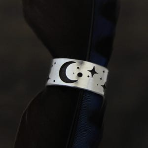May include: A silver-colored ring with a celestial design. The ring features a crescent moon, stars, and small dots. The ring is wrapped around a dark, textured object, creating a contrast between the shiny metal and the dark background.