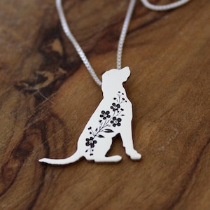 May include: A silver-colored pendant necklace featuring a dog silhouette with floral detailing. The dog is sitting, and the flowers are black. The necklace chain is also silver and is on a wooden surface.
