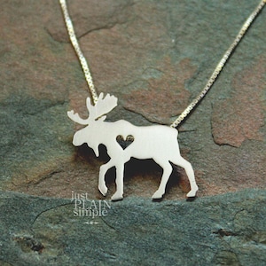 May include: A silver-toned necklace featuring a moose-shaped pendant with a small heart-shaped cutout in the centre of the body. The pendant is attached to a thin, delicate box chain. The text "just PLAIN simple" is visible in the lower left corner.