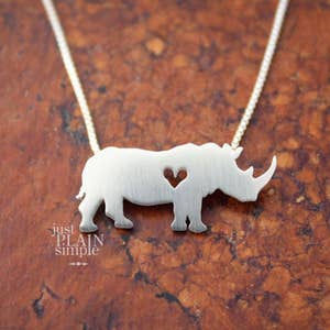 May include: A silver necklace with a rhinoceros pendant featuring a heart cut-out. The pendant is on a silver chain. The text "just PLAIN simple" is visible below the pendant.