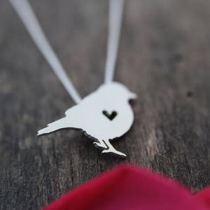 May include: A silver necklace featuring a bird pendant. The bird has a heart-shaped cutout in its chest.