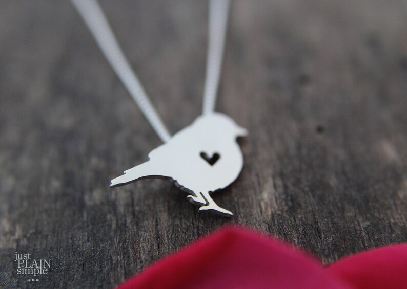 May include: A silver necklace featuring a bird pendant. The bird has a heart-shaped cutout in its chest.