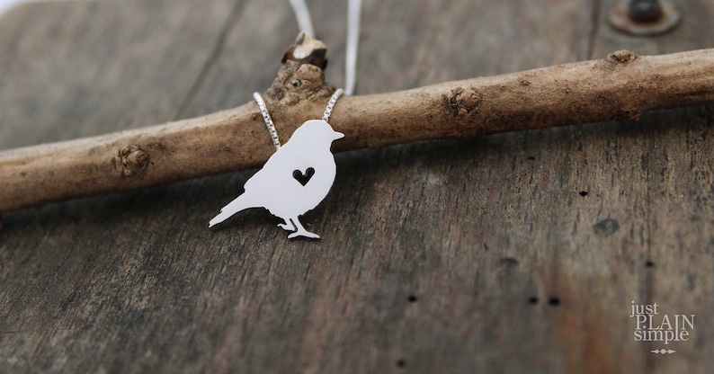 May include: A silver necklace featuring a bird pendant. The bird has a heart cut out of its chest. The necklace is hanging on a branch.