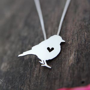 May include: A silver necklace featuring a bird pendant. The bird has a small heart cut out of its chest.