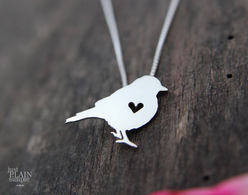 May include: A silver necklace featuring a bird pendant. The bird has a small heart cut out of its chest.