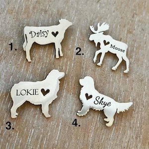 May include: Four silver metal pet tags with different animal silhouettes and names. The tags are numbered 1 through 4. The tags feature a cow with the name Daisy, a moose with the name Moose, a dog with the name Lokie, and a dog with the name Skye.