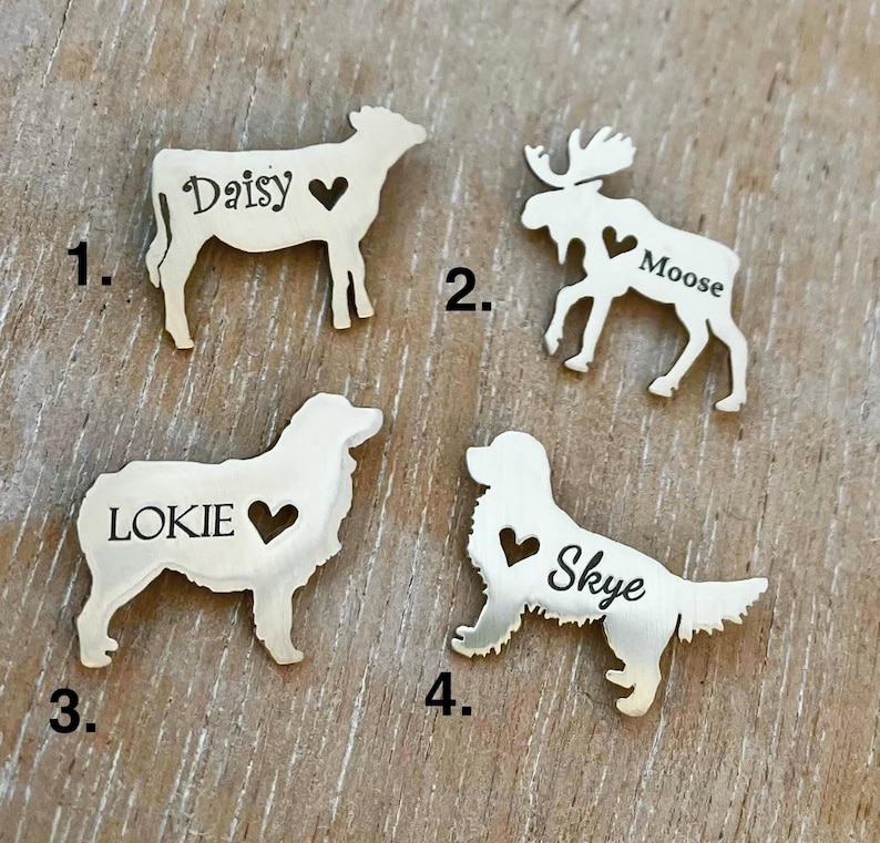 May include: Four silver metal pet tags with different animal silhouettes and names. The tags are numbered 1 through 4. The tags feature a cow with the name Daisy, a moose with the name Moose, a dog with the name Lokie, and a dog with the name Skye.