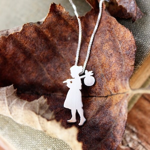 May include: A silver pendant necklace featuring a silhouette of a person carrying a bundle and a staff. The pendant hangs on a delicate chain, set against a backdrop of a dried leaf and a book.