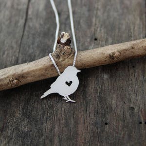 May include: A silver necklace with a bird pendant featuring a heart cutout. The bird is perched on a branch.