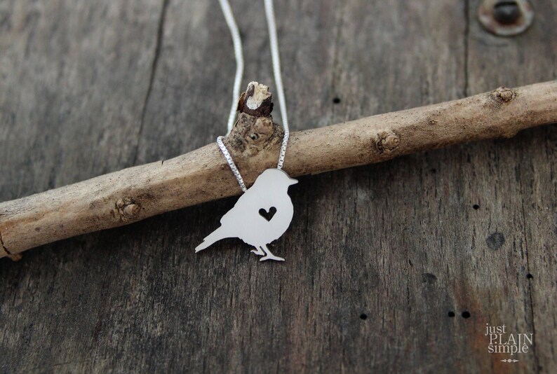 May include: A silver necklace with a bird pendant featuring a heart cutout. The bird is perched on a branch.
