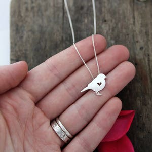 May include: A silver necklace featuring a dainty bird pendant. The bird has a small heart cut out of its chest.