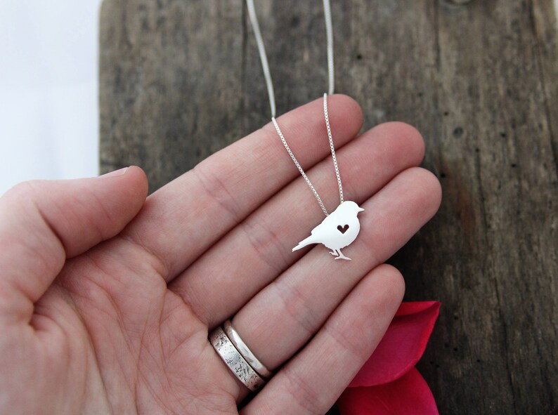 May include: A silver necklace featuring a dainty bird pendant. The bird has a small heart cut out of its chest.