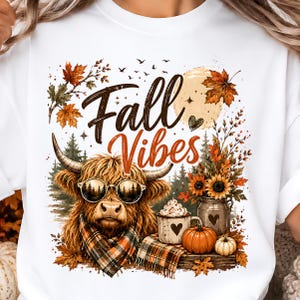 Può includere: Una t-shirt bianca con la grafica di una mucca Highland che indossa occhiali da sole e una sciarpa a quadri. Il design include il testo Fall Vibes, una luna, foglie autunnali, una tazza con panna, una brocca di metallo e due zucche arancioni.