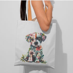 May include: White tote bag featuring a colorful illustration of a puppy adorned with floral patterns. The puppy has a black and white face and is surrounded by flowers in shades of pink, red, and green. The bag is held by a person.
