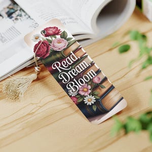 May include: A decorative bookmark featuring a floral design with red and pink roses, daisies, and the text "Read, Dream, Bloom." The bookmark has a tassel and is placed on a wooden surface next to an open book.